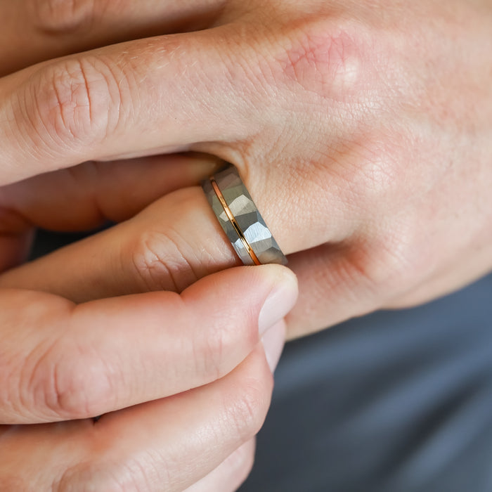 Close-up of a hand wearing The Sheridan, an 8mm men's textured silver Tungsten Carbide ring with a thin gold stripe, as another hand gently touches it against a blurred background.