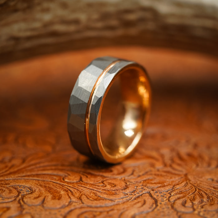 The Sheridan, an 8mm men's ring crafted from durable Tungsten Carbide, features a textured, faceted gray band and a luxurious Rose Gold interior. Resting on an ornate, embossed brown surface with a blurred background, this striking ring blends modern elegance with timeless style.