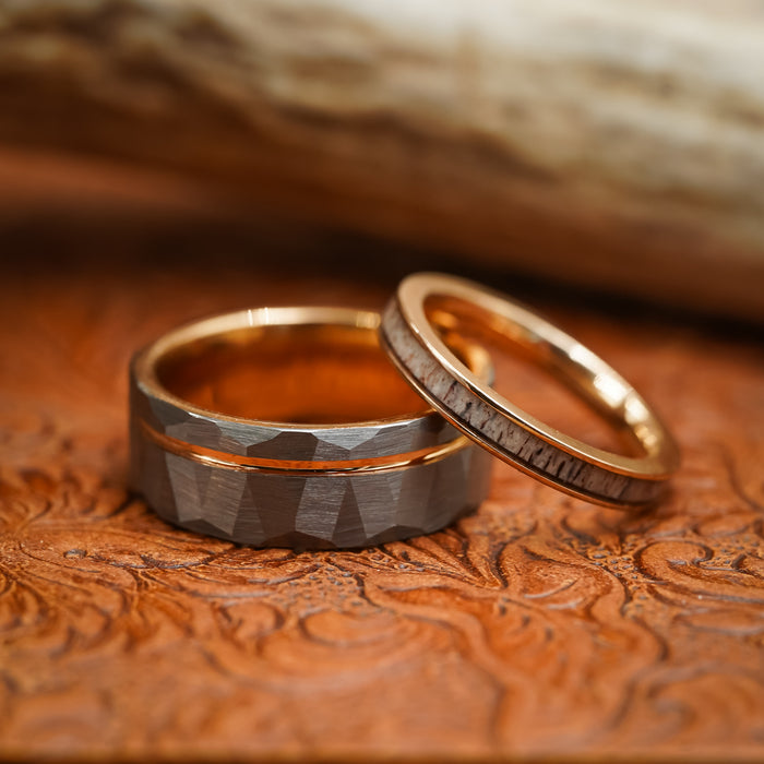 Two rings rest on a textured brown surface: one is The Sheridan, a wide men's 8mm Tungsten Carbide ring with faceted edges and a striking Rose Gold interior band, while the other is thin, gold, and inlaid with a light-colored material.