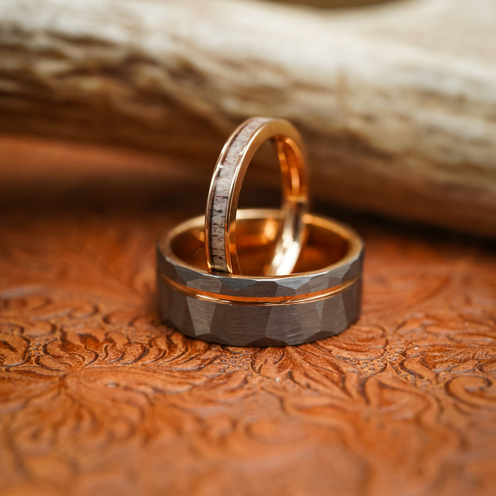 Two rings rest on a tooled leather surface: one gold with a light stone inlay, and the other is The Sheridan—a wider men's 8mm Tungsten Carbide ring featuring a dark matte hammered texture, copper accent, and a subtle hint of Rose Gold interior. A blurred beige background is visible.