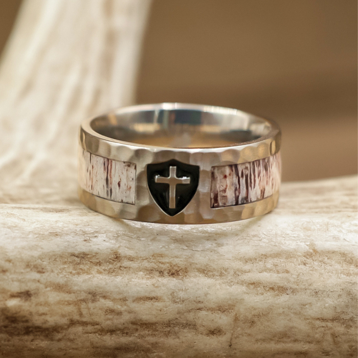The Shield of Faith, a silver Christian wedding band adorned with a black shield and cross emblem, showcases rustic wood-like inlays and a subtle hammered finish as it rests on a textured, light-colored antler.
