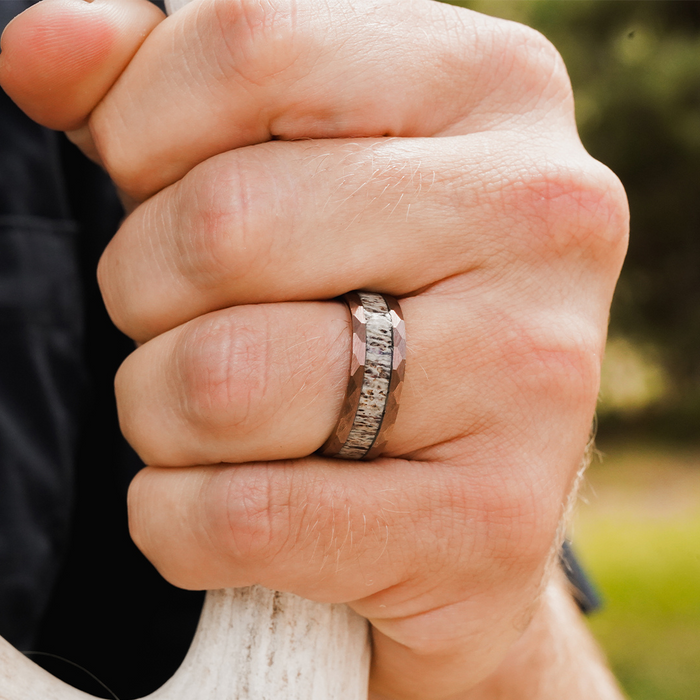 A close-up of a hand gripping a white object, wearing The Sierra Nevada ring—a metallic band featuring a hammered edge and textured pattern. The blurred background reveals hints of greenery.