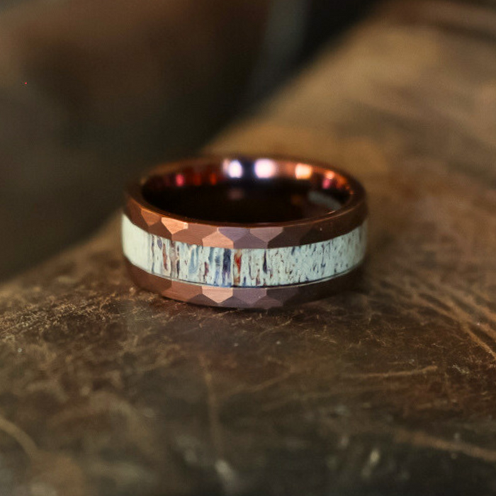 A close-up of The Sierra Nevada Ring, featuring a faceted metallic reddish-brown Tungsten Carbide band with a light, textured antler inlay, resting on a worn brown surface.