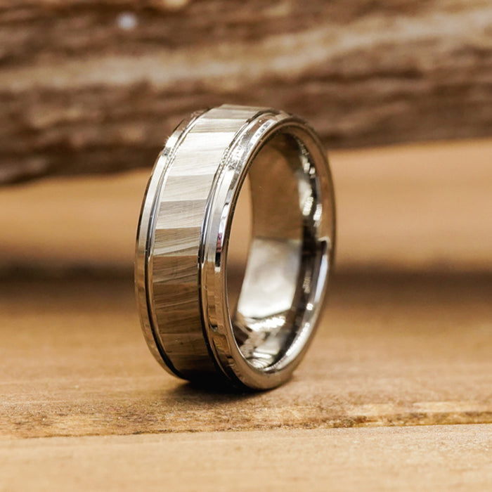 The Silverton, a rugged men's ring crafted from silver metal with a brushed center band and polished edges, stands upright on a wooden surface against a blurred wooden background.