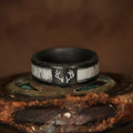 The Stag is a distinctive men’s wedding band crafted from black forged carbon fiber, featuring a striking silver deer antler design and a white inlay. Set against a rustic, weathered metal surface with a blurred brown background, this ring blends rugged elegance with modern style.