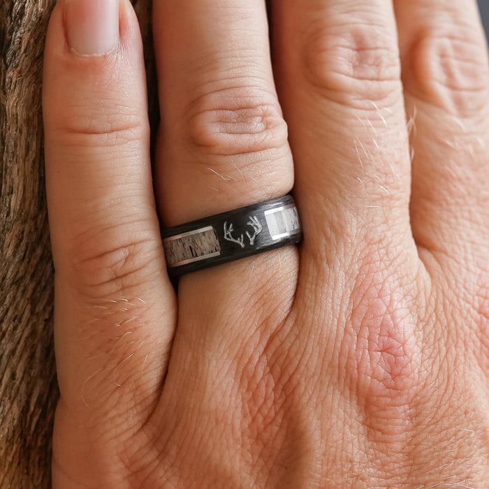 A close-up of a hand wearing The Stag—a black forged carbon fiber ring featuring antler designs and light-colored inlays—resting against a textured surface. This distinctive men's wedding band seamlessly blends rugged character with refined accents, making it a truly unique statement piece.