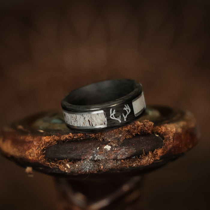 Introducing The Stag: a striking men's wedding band crafted from black forged carbon fiber, featuring luminous inlay bands and a distinctive white deer antler inlay design. This unique ring rests elegantly on a rustic, textured wooden surface with a blurred brown background, showcasing its exceptional craftsmanship and bold presence.