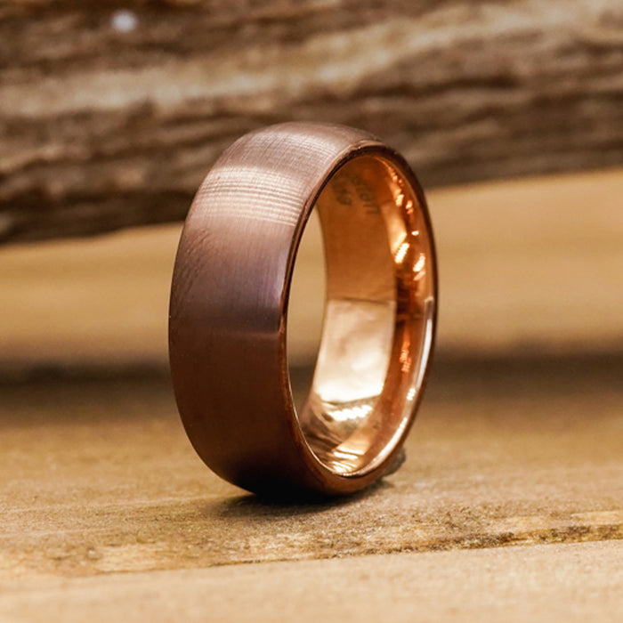 A close-up of The Taos, an 8mm matte Brown Rose Gold ring expertly crafted from durable Tungsten Carbide, standing upright on a wooden surface against a softly blurred wooden background.