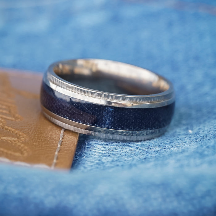 The Ty, a silver titanium ring featuring a dark blue inlay, is displayed on light blue denim fabric beside a brown leather patch, creating the perfect look for a men's rugged band.