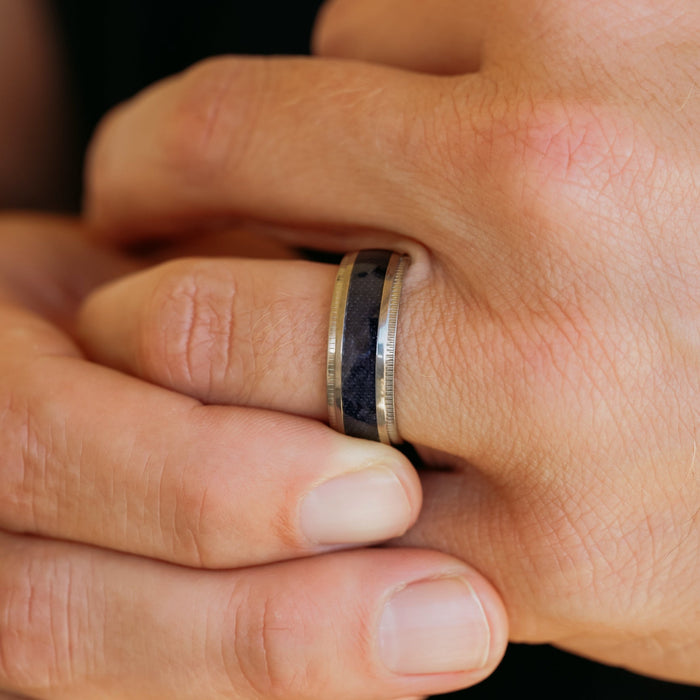 A close-up of a hand wearing The Ty—a men's rugged silver ring featuring a dark, patterned inlay reminiscent of antler rings. Another hand gently clasps the finger adorned with The Ty, emphasizing its distinctive design.