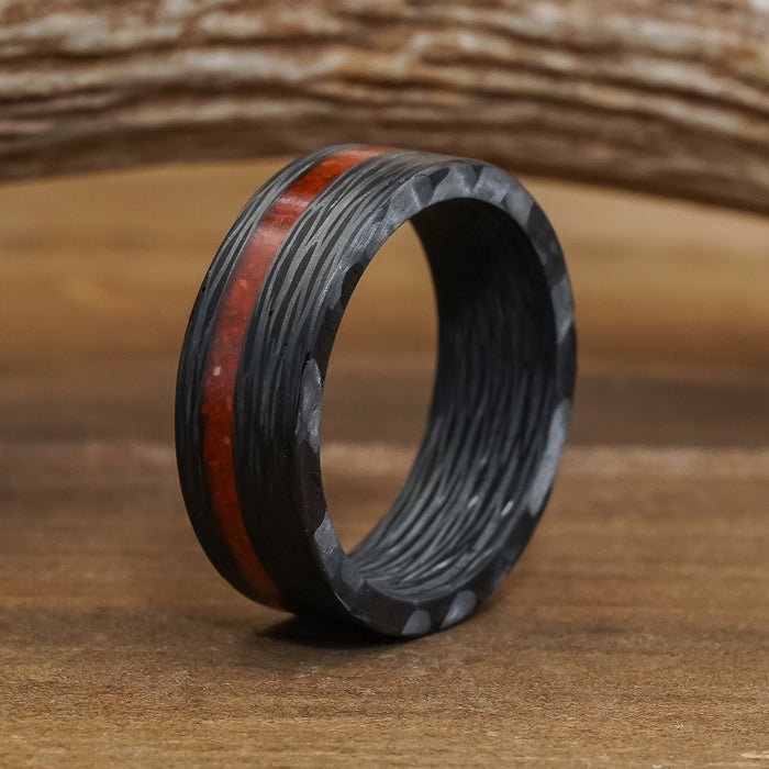 The Valley Of Fire, an 8mm men's carbon fiber ring featuring a textured surface and a striking reddish-brown inlay, stands upright on a wooden surface with a blurred wooden object in the background.