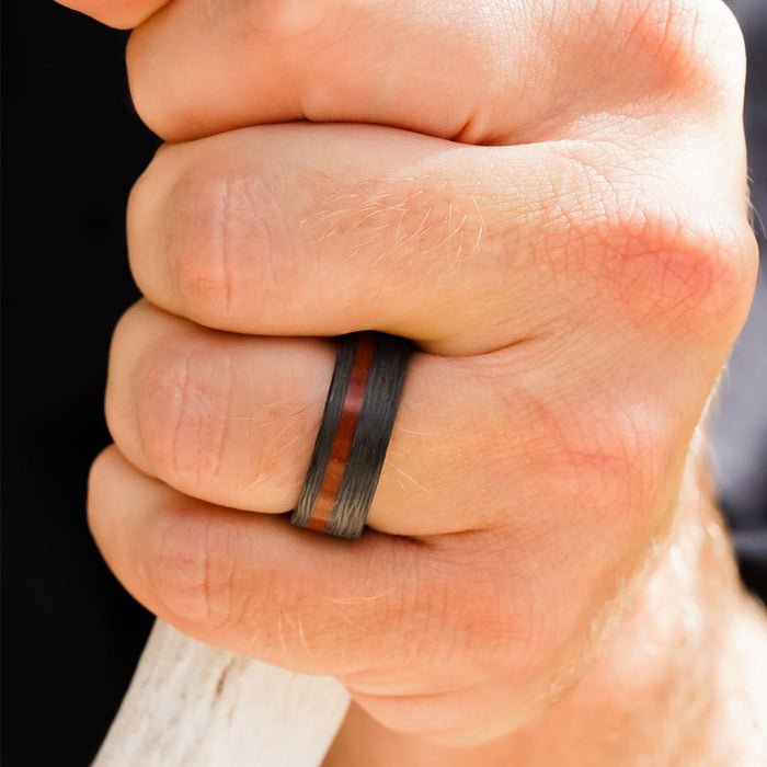 A close-up of a hand gripping a white object, wearing The Valley Of Fire men's 8mm ring—a striking piece featuring a bold reddish-brown stripe down the center reminiscent of red jasper gemstone.