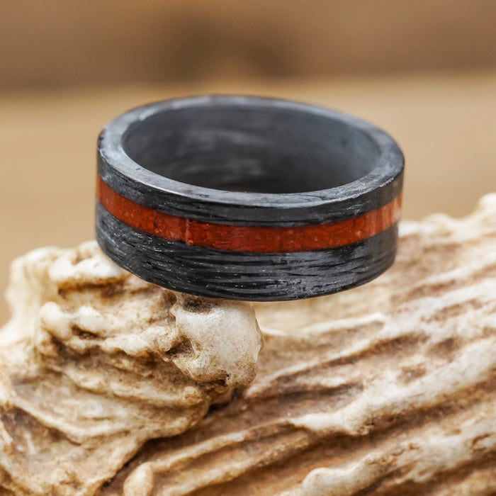 The Valley Of Fire men’s 8mm ring showcases a textured matte black finish complemented by a bold red-brown stripe inspired by the vibrant hues of red jasper gemstone, elegantly presented atop a rugged piece of natural beige stone.