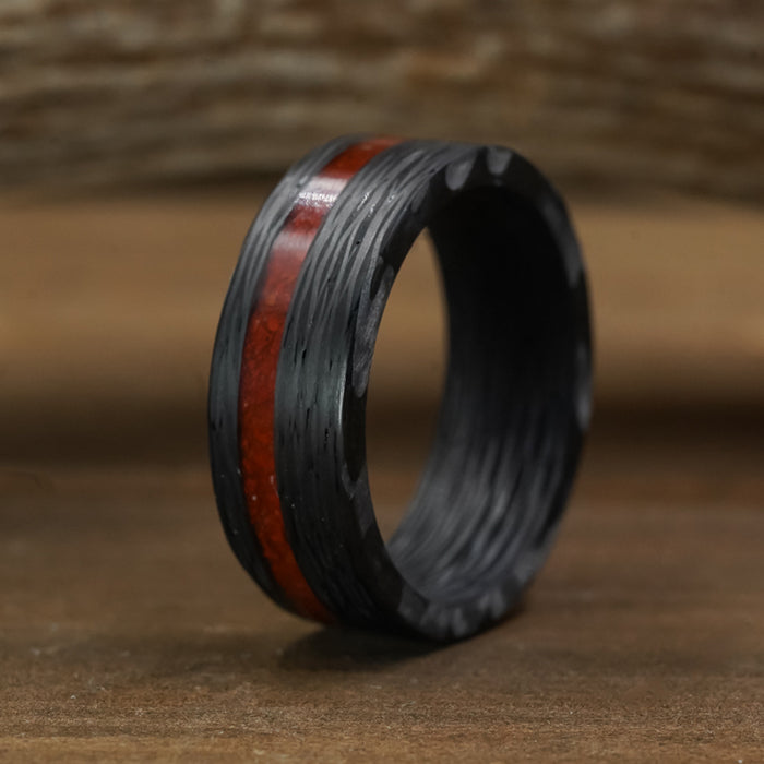 The Valley Of Fire men's 8mm ring showcases a black textured band accented by a glossy red inlay running through the center, displayed upright on a wooden surface with a softly blurred wooden background.