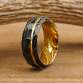 The Vogel ring—a black hammered band featuring a jagged gold stripe and a brilliant yellow-gold interior—stands upright on a wooden surface, its distinctive texture and design creating a stunning contrast against the softly blurred, rustic background.
