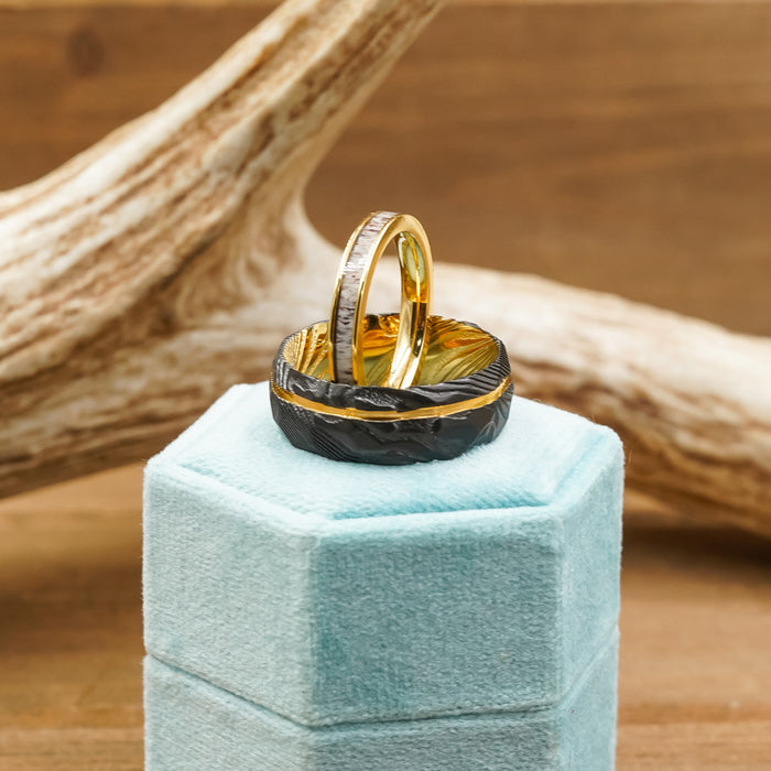 Two "The Vogel" rings—one in gold and the other in a dark finish with a textured pattern reminiscent of Damascus steel—are displayed side by side on a light blue velvet hexagonal ring box, with a piece of wood featuring a natural, light finish in the background.