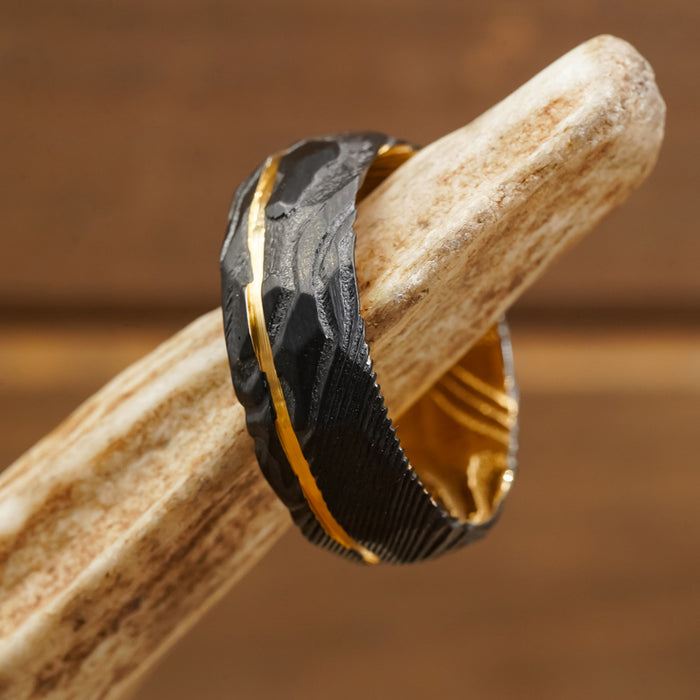 The Vogel, a textured black hammered ring featuring a yellow-gold interior and a twisted design, is displayed on a smooth, light-colored antler against a brown background.