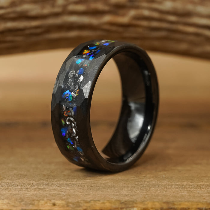 Introducing The Wickenburg: a black Tungsten Carbide ring with a faceted surface, adorned with vibrant galaxy opal inlays that evoke distant stars, elegantly displayed on a wooden surface against a softly blurred wooden background.