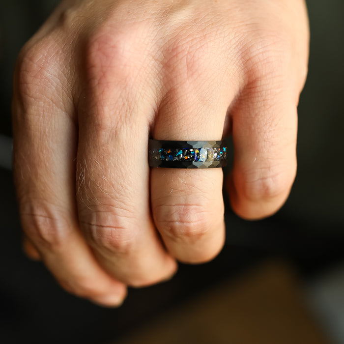 A close-up of a hand wearing The Wickenburg, a matte black Tungsten Carbide ring with blue and white stones set in a textured band on the ring finger.