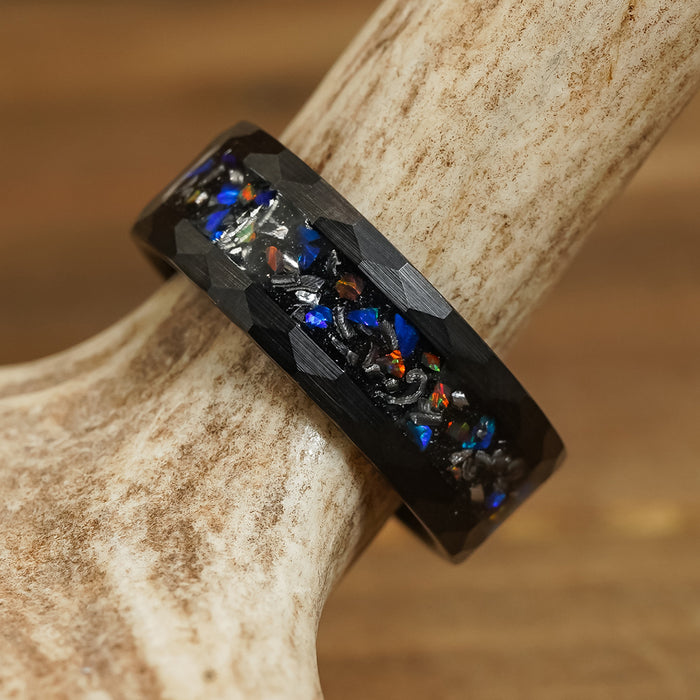 The Wickenburg, a black Tungsten Carbide ring featuring a vibrant inlay of iridescent stones reminiscent of galaxy opal, is displayed resting on a light-tan antler with a softly blurred brown background.