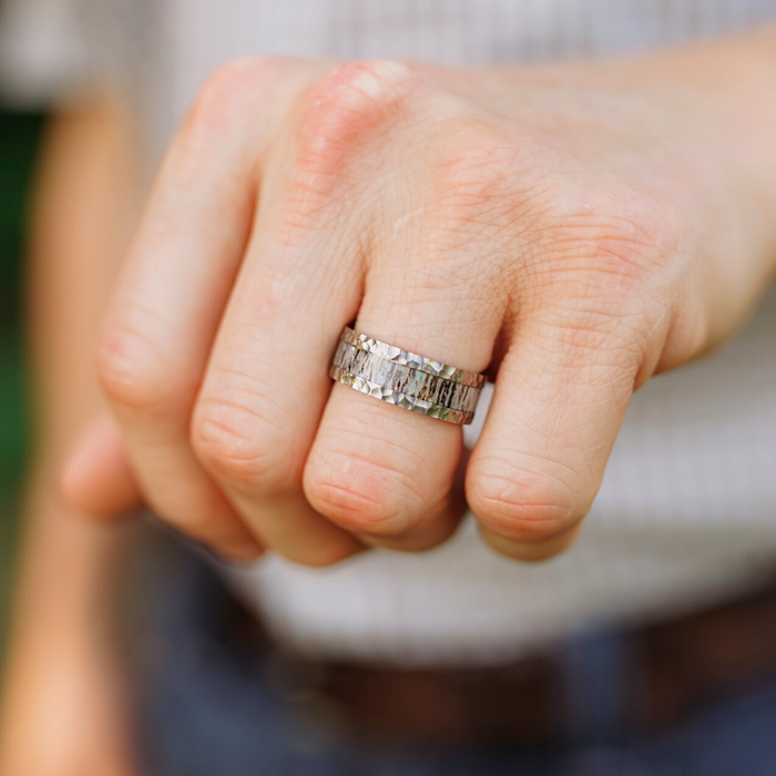 A close-up of a hand wearing The Wilder Ranch men's hammered ring, featuring a textured geometric design, held in a loose fist. The person is dressed in a striped shirt and a dark belt.