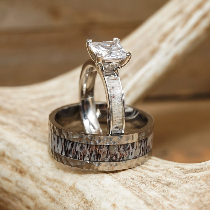 Two silver rings from The Wilder Ranch collection rest on a textured antler. One is an antler wedding band with a square-cut clear gemstone, while the other is a rugged men’s hammered ring in brown and silver tones. The softly blurred wood in the background adds warmth to the scene.