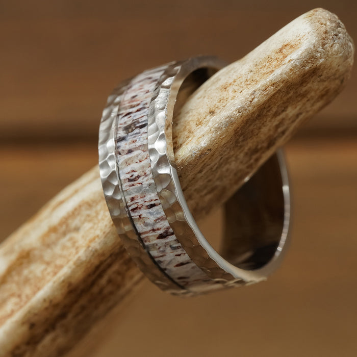 The Wilder Ranch, a rugged titanium ring featuring a hammered texture and a central inlay reminiscent of antler or wood, is showcased atop the tip of a light brown antler with a softly blurred brown background.