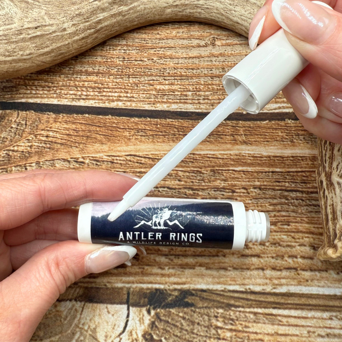 A hand holds a tube labeled Travel Jewelry Cleaner while another hand opens it, revealing a white brush applicator. The scene showcases the ease of using this non-toxic jewelry cleaner, set against antler decor and a textured wood surface.
