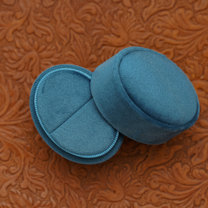 Two blue Velvet Oval Ring Boxes, ideal for proposal photo shoots or wedding photography, rest on an embossed brown leather surface with a delicate floral pattern—one box open and the other closed.