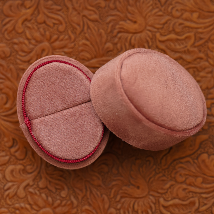 Two pink Velvet Oval Ring Boxes rest on an embossed brown surface with a floral pattern. Perfect for proposal photo shoots or wedding photography, one box is open to reveal its elegant inner lining while the other remains closed.