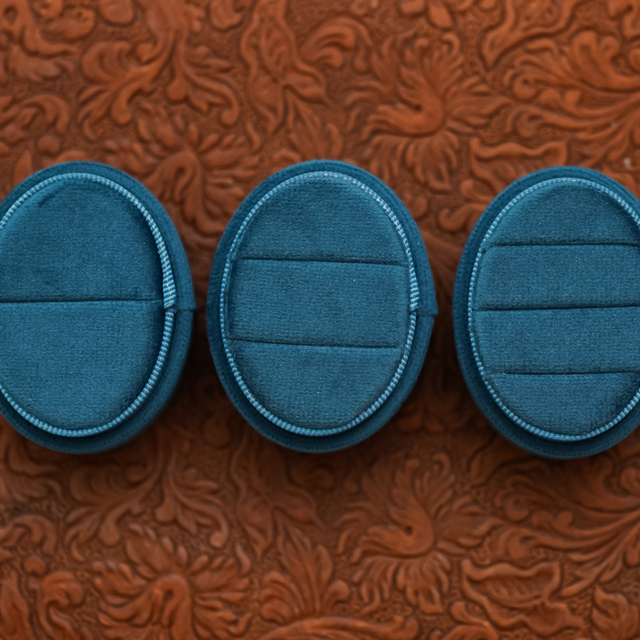 Three empty Velvet Oval Ring Boxes in blue are arranged in a row on a textured brown surface with a floral pattern, making them ideal props for wedding photography or proposal photo shoots.